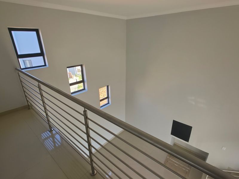 A bright, modern upper-level landing featuring a sleek stainless steel balustrade, tiled flooring, and multiple windows overlooking the lower level.