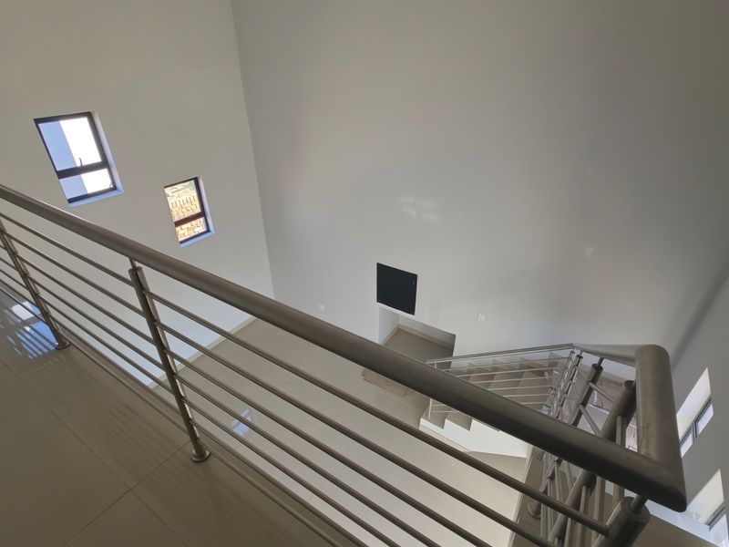 A bright, double-volume landing area featuring modern stainless steel balustrades, tiled flooring, and a staircase leading to the lower level.