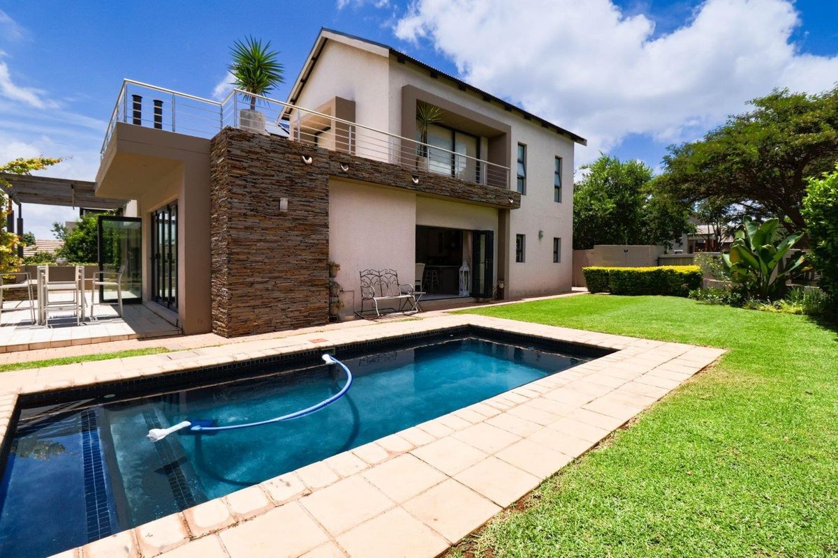 Rear view of a modern double-story house featuring a swimming pool, landscaped garden, tiled patio, and an upper-level balcony with stone cladding accents.