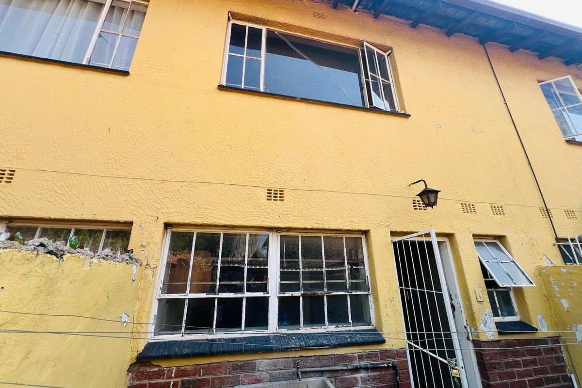Rear exterior view of the apartment building showing yellow textured facade, steel-framed windows, security gate, and a concrete utility sink.