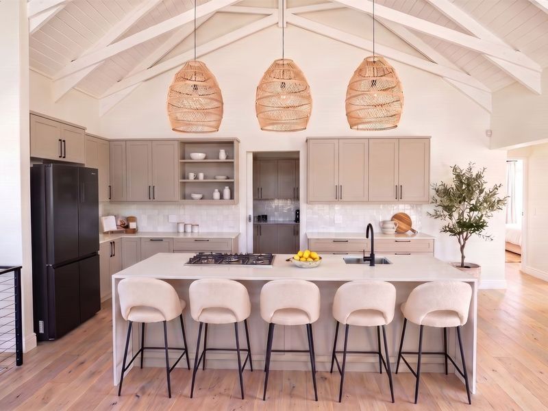 Modern open-plan kitchen featuring a large central island with gas cooktop, shaker cabinetry, vaulted ceilings with exposed rafters, and light hardwood flooring.