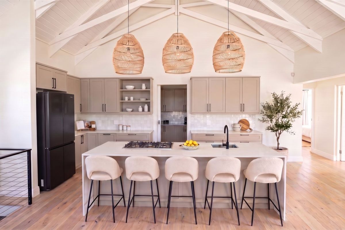 Modern open-plan kitchen featuring a large central island with gas cooktop, shaker cabinetry, vaulted ceilings with exposed rafters, and light hardwood flooring.