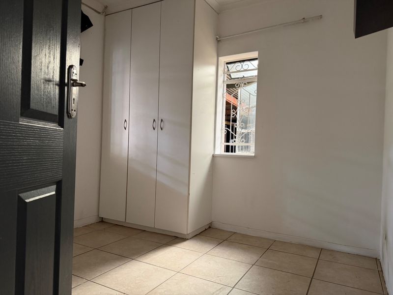 A clean, empty bedroom featuring neutral tiled flooring, a built-in wardrobe, and a window fitted with a security gate.