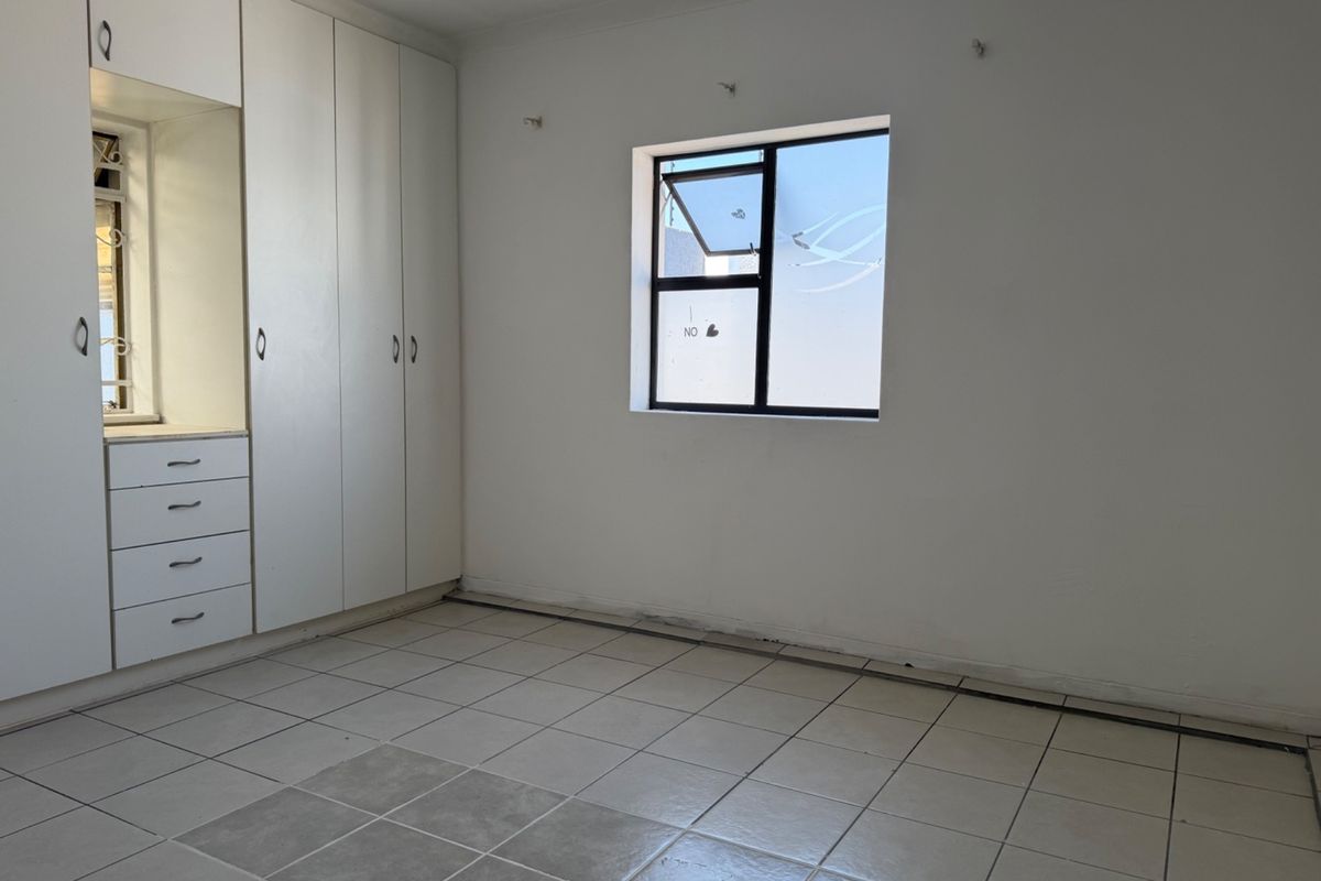 An empty bedroom featuring white built-in cupboards, tiled flooring, and a single window with an aluminium frame.
