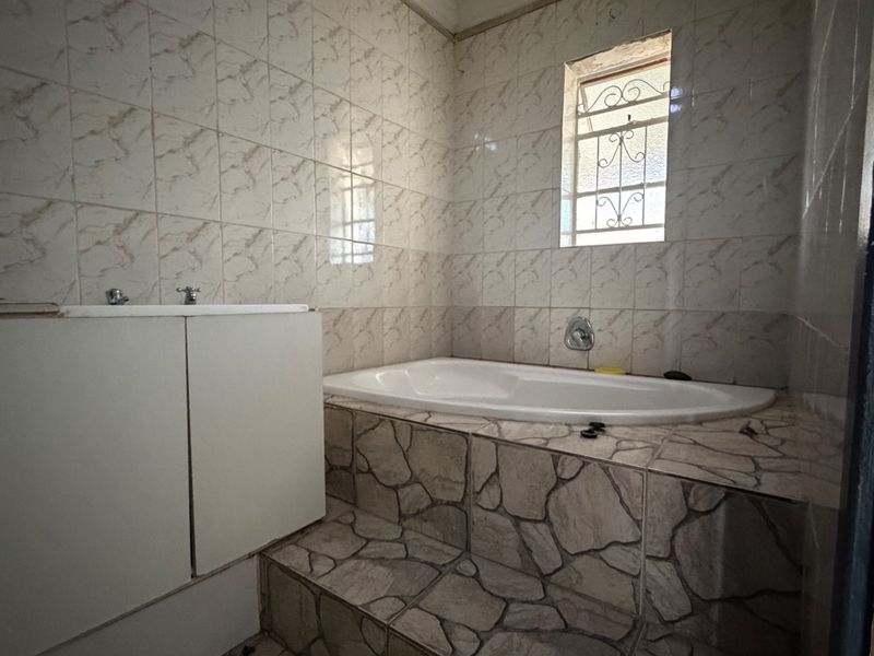 A dated bathroom featuring a built-in bathtub with stone-effect tiled surround and wall tiles, complemented by a security-grille window.