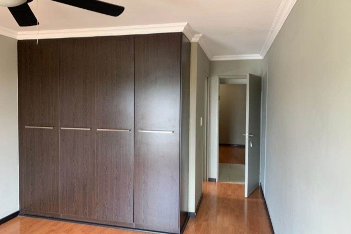 A clean bedroom featuring dark wood built-in cupboards, a ceiling fan, and warm-toned laminate flooring, leading into an adjacent hallway.
