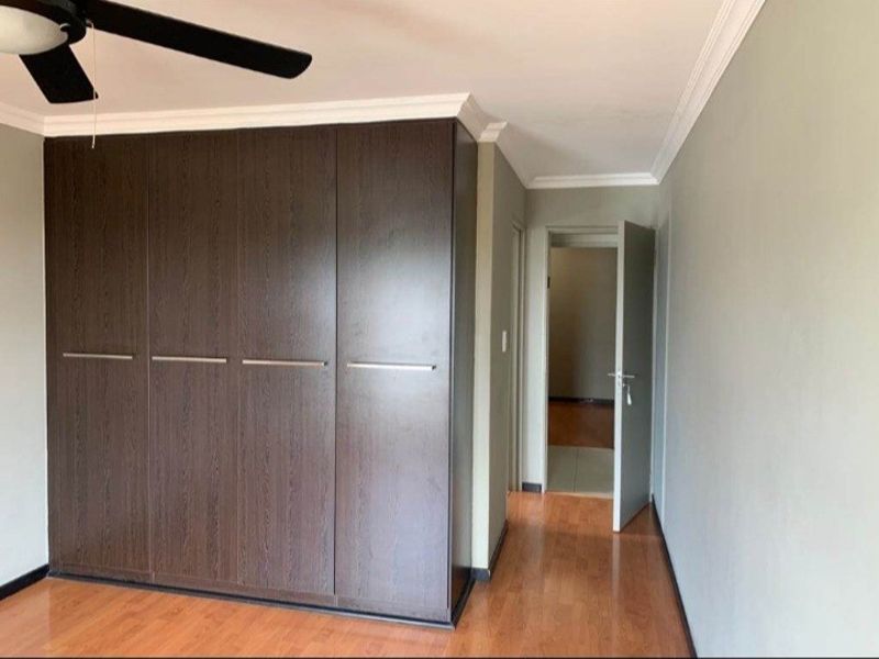 A clean bedroom featuring dark wood built-in cupboards, a ceiling fan, and warm-toned laminate flooring, leading into an adjacent hallway.