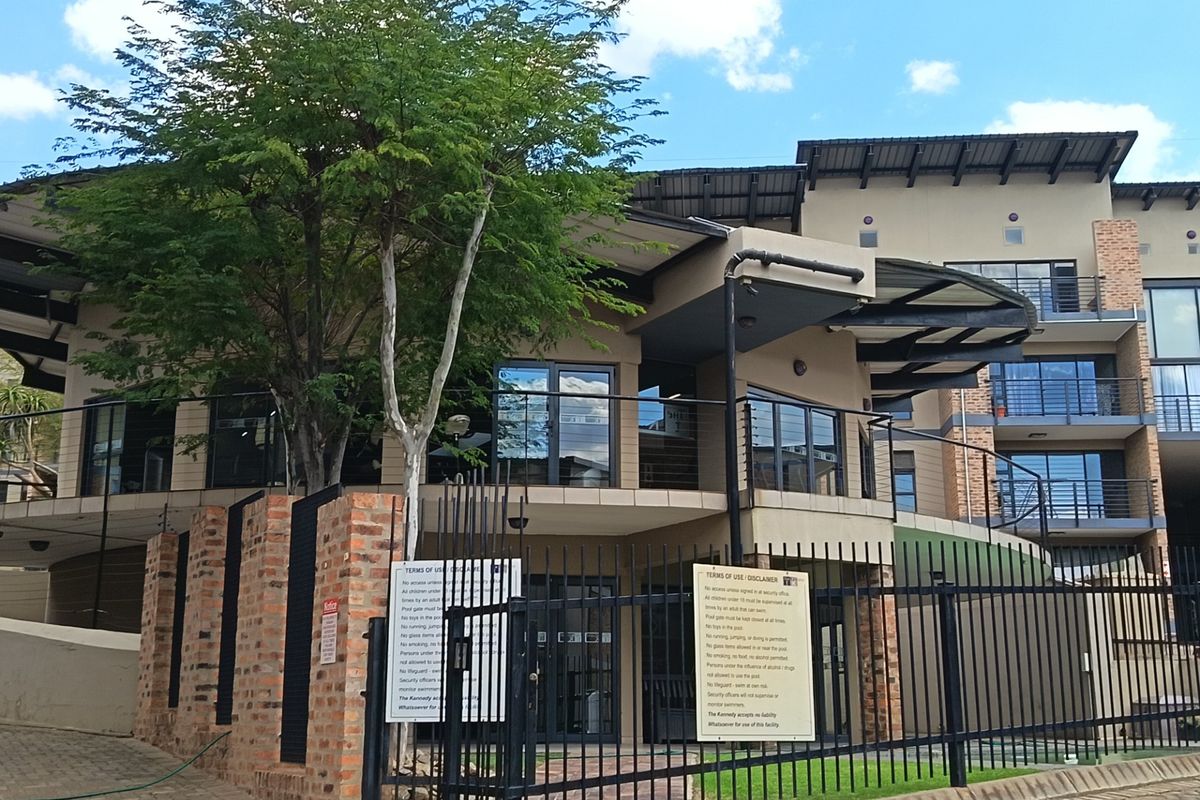 Exterior view of a modern apartment complex featuring brick boundary walls, metal security fencing, and multi-level balconies with sliding glass doors.
