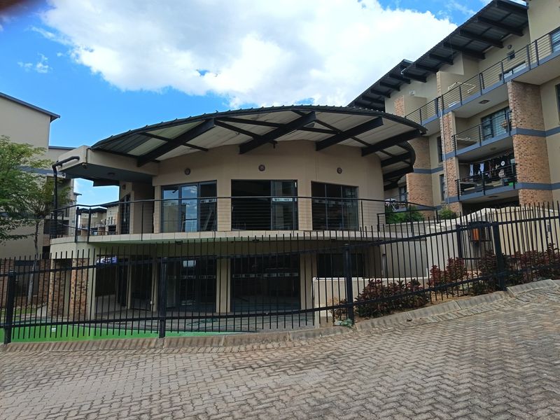 A modern clubhouse building with a distinctive curved roof design, featuring steel balustrades and adjacent multi-story apartment units within a secure complex.