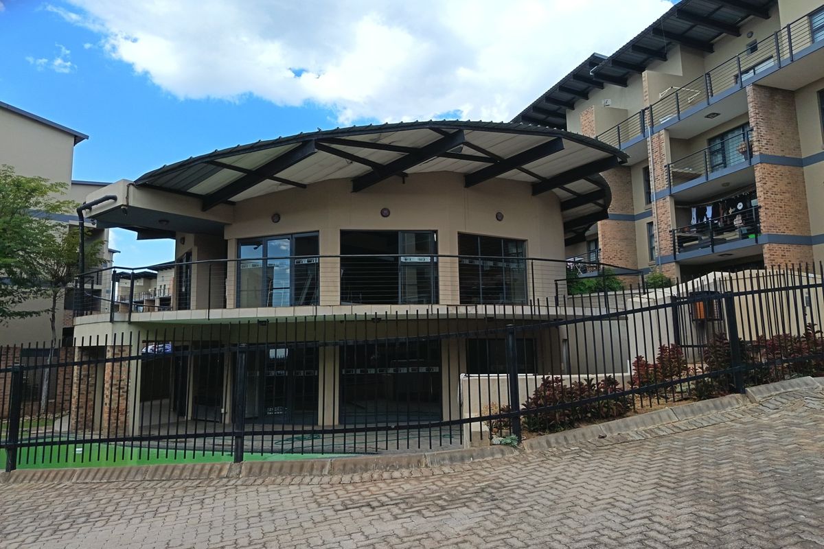 A modern clubhouse building with a distinctive curved roof design, featuring steel balustrades and adjacent multi-story apartment units within a secure complex.