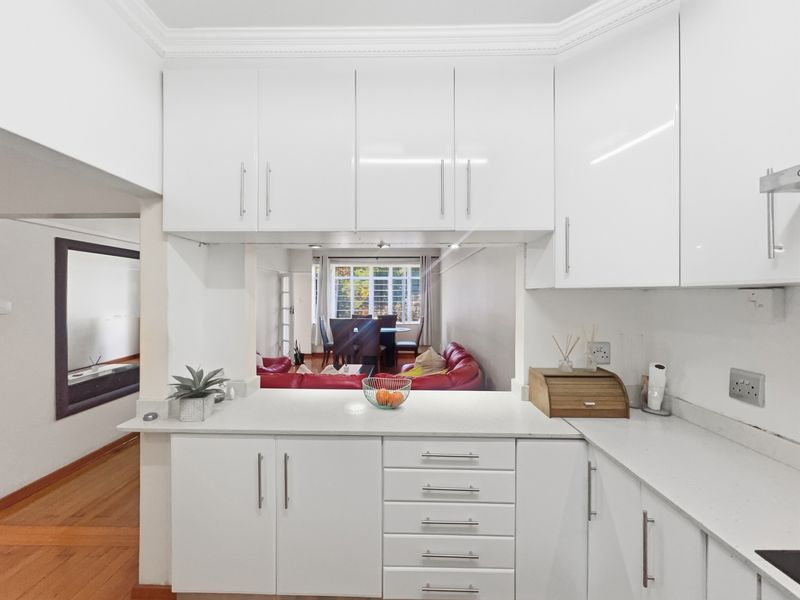 Modern kitchen featuring white gloss cabinetry, stone countertops, and elegant coving, with a convenient pass-through window overlooking the adjacent living and dining area.