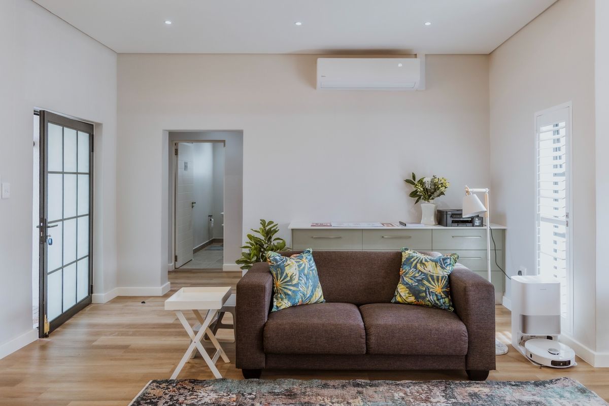 A bright and modern living room featuring light laminate flooring, recessed downlights, a wall-mounted air conditioning unit, and elegant shutter blinds.