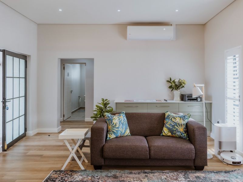 A bright and modern living room featuring light laminate flooring, recessed downlights, a wall-mounted air conditioning unit, and elegant shutter blinds.