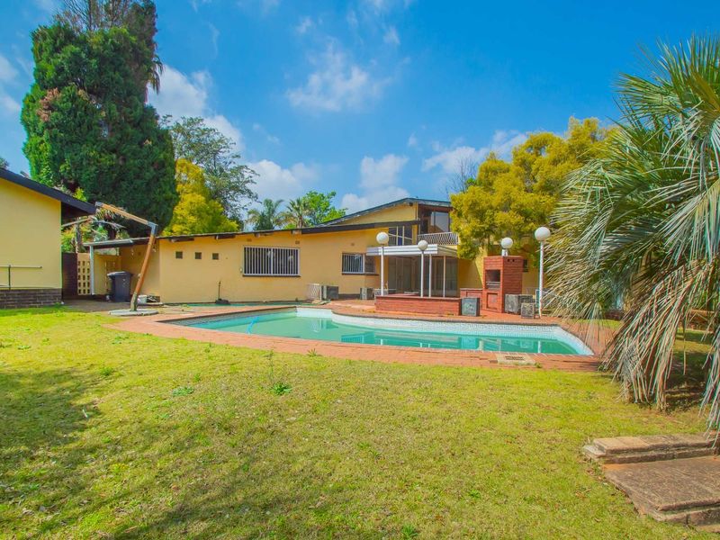 Rear exterior view featuring a swimming pool, brick-built braai area, and established garden space with paved surrounds.