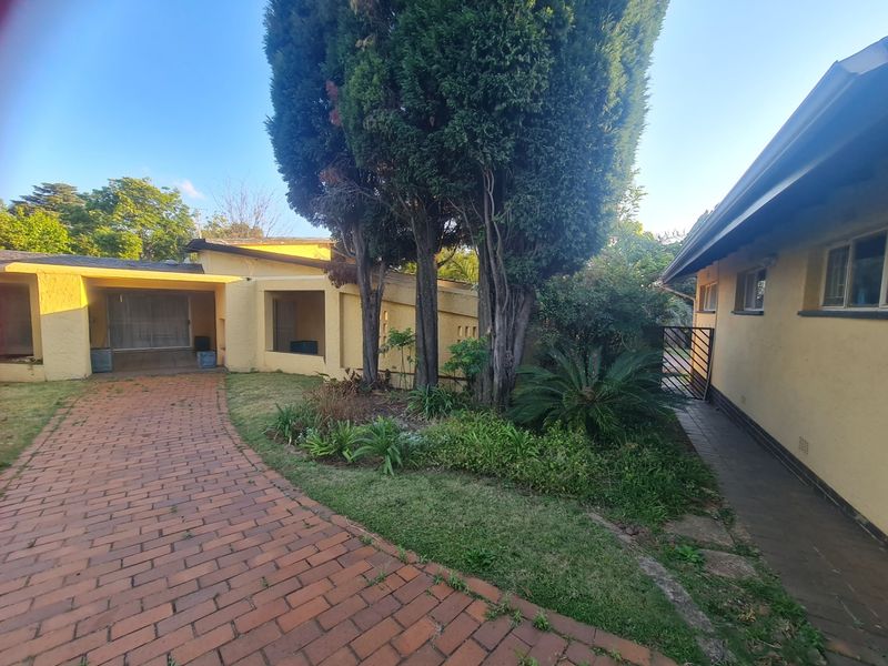 A view of the rear exterior featuring a brick paved driveway, a covered patio area, and a mature landscaped garden with established trees.