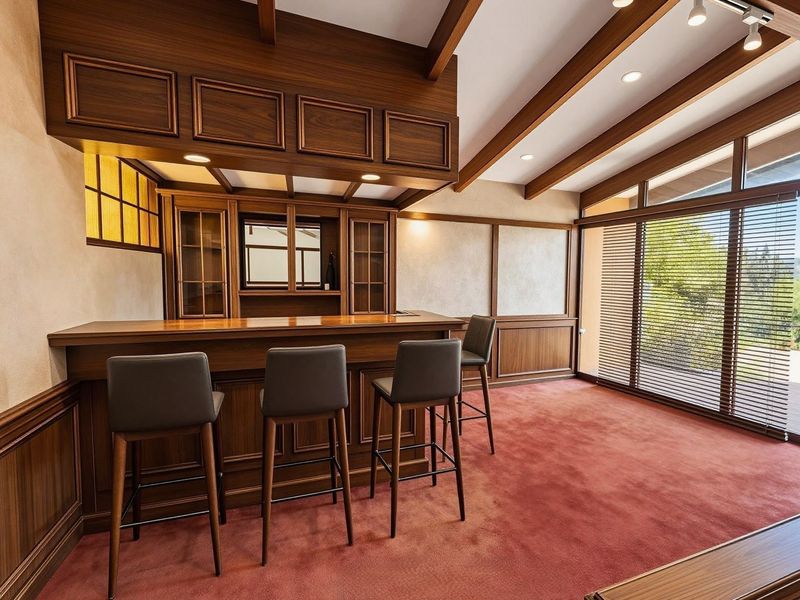 A well-appointed home bar featuring custom wood cabinetry, a built-in counter, exposed ceiling beams, and large windows providing natural light and garden views.