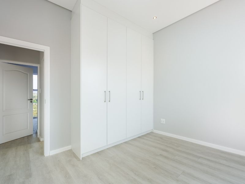 A bright, modern bedroom featuring neutral grey walls, light wood-look laminate flooring, and a large, clean-lined white built-in wardrobe.