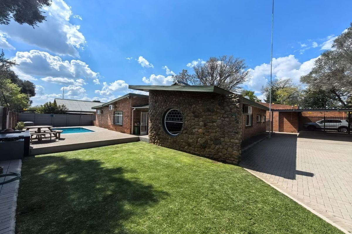 Rear exterior view featuring a swimming pool, manicured lawn, stone-clad feature wall with circular window, and a paved driveway leading to a carport.