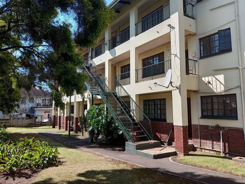 Rear view of a multi-story apartment building featuring external metal staircases, balconies with railings, and a shared garden area.