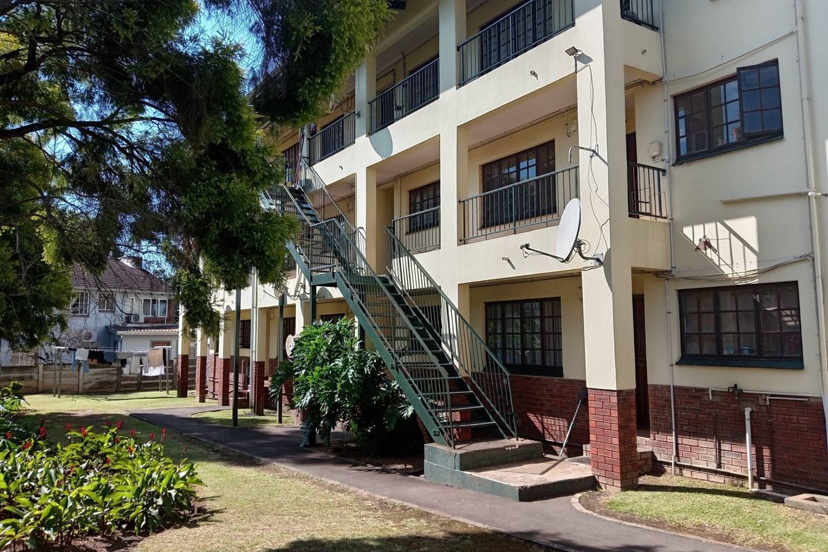 Rear view of a multi-story apartment building featuring external metal staircases, balconies with railings, and a shared garden area.