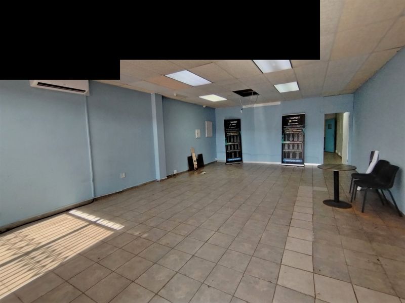 An empty, open-plan commercial or living space featuring ceramic tile flooring, a drop ceiling with recessed lighting, and a wall-mounted air conditioning unit.