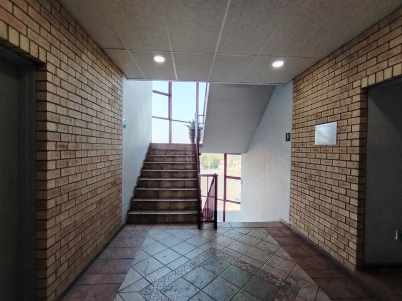 A spacious hallway featuring exposed brick walls, tiled flooring, and a staircase with a metal balustrade, illuminated by recessed ceiling lighting.