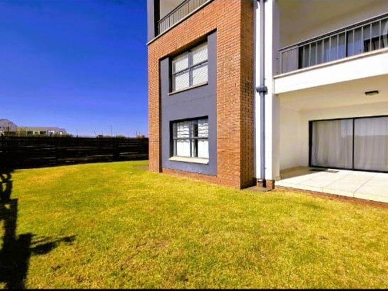 Rear view of the apartment building featuring a private garden, tiled patio area, and sliding glass door access to the interior.