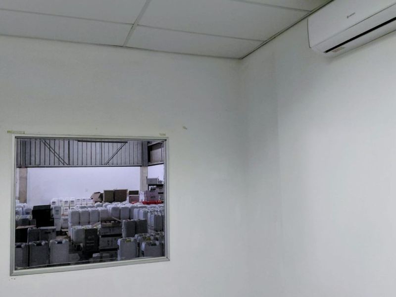 A functional office space featuring a suspended ceiling, wall-mounted air conditioning unit, and an internal window overlooking the warehouse area.