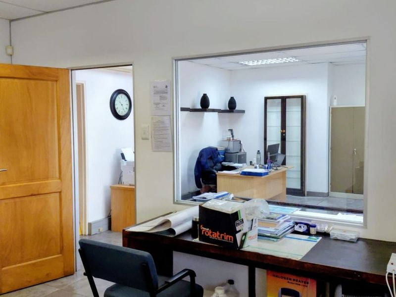 An office space featuring a large glass partition, tiled flooring, and a wooden door, providing a functional workspace with clear visibility between rooms.