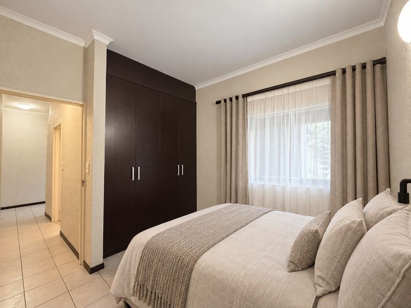 A well-maintained bedroom featuring dark wood built-in cupboards, neutral tiled flooring, and a window with fitted curtain rods.