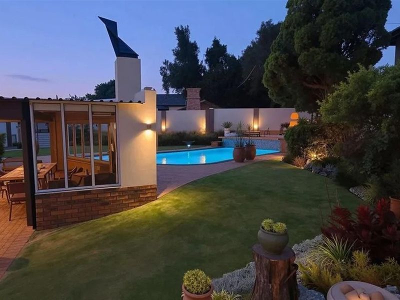 A beautifully landscaped backyard featuring a swimming pool, covered patio area with an outdoor fireplace, and ambient evening lighting.