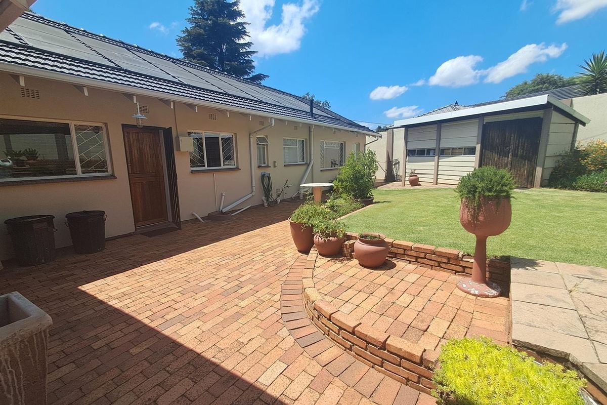 Rear exterior view featuring a brick-paved patio, landscaped lawn, retaining wall, and a detached garage with a tiled roof and solar panels.