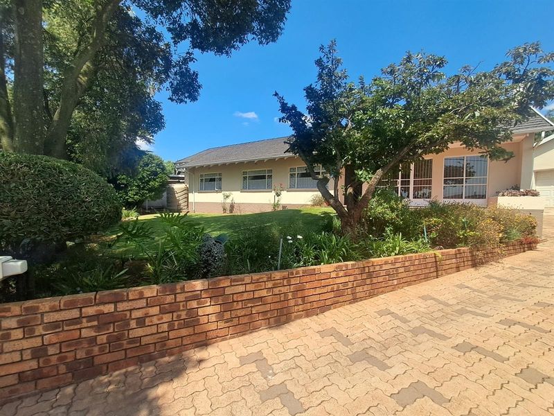 Front view of a single-story house featuring a paved driveway, a brick retaining wall, and a well-maintained garden with mature trees.