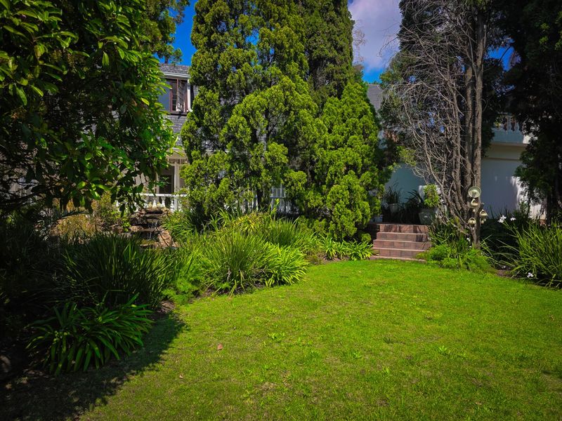 A lush, well-maintained garden featuring a green lawn, mature trees, a small water feature, and brick steps leading toward the house.