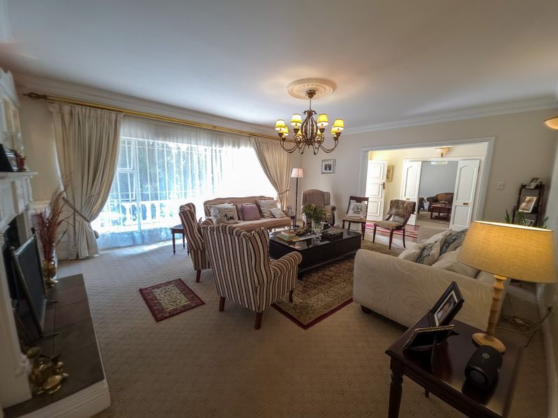 Spacious living room featuring a fireplace, decorative ceiling rose with chandelier, and large windows allowing natural light, with double doors leading to adjacent rooms.