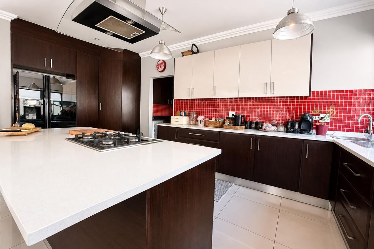 Modern kitchen featuring a central island with gas hob, dark wood cabinetry, contrasting white upper cabinets, stone countertops, and a vibrant red tiled backsplash.