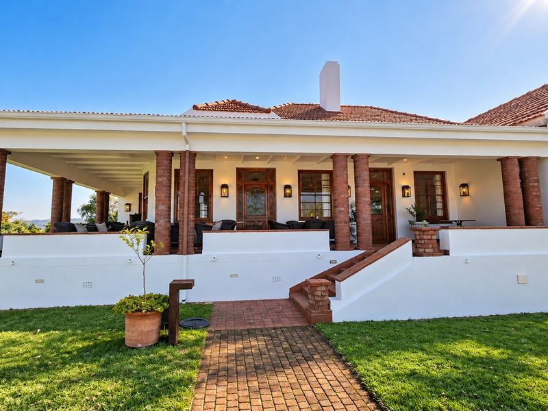 A well-maintained rear exterior view featuring a spacious covered patio with brick columns, terracotta roof tiles, and a paved walkway leading to the entrance.