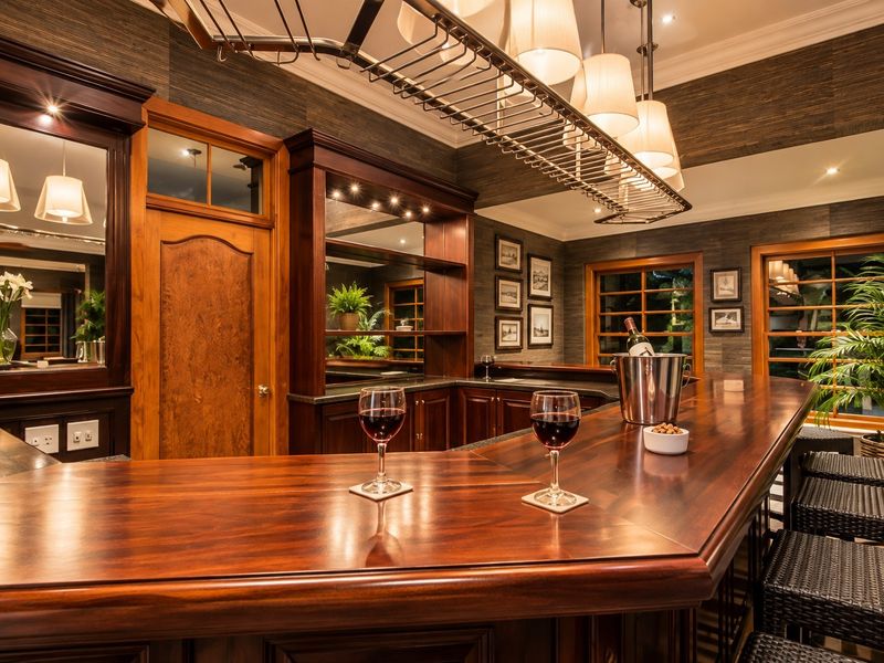 A sophisticated built-in home bar featuring rich timber cabinetry, a polished wooden counter, mirrored back bar, and elegant overhead lighting.