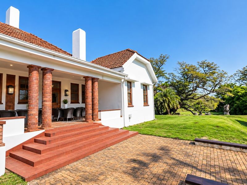 A well-maintained rear exterior view featuring a covered patio with brick columns, tiled steps, and a spacious garden area with tennis court fencing.