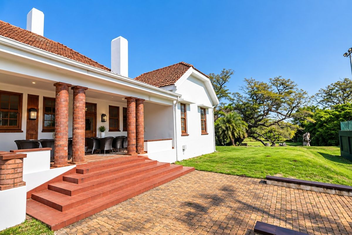A well-maintained rear exterior view featuring a covered patio with brick columns, tiled steps, and a spacious garden area with tennis court fencing.