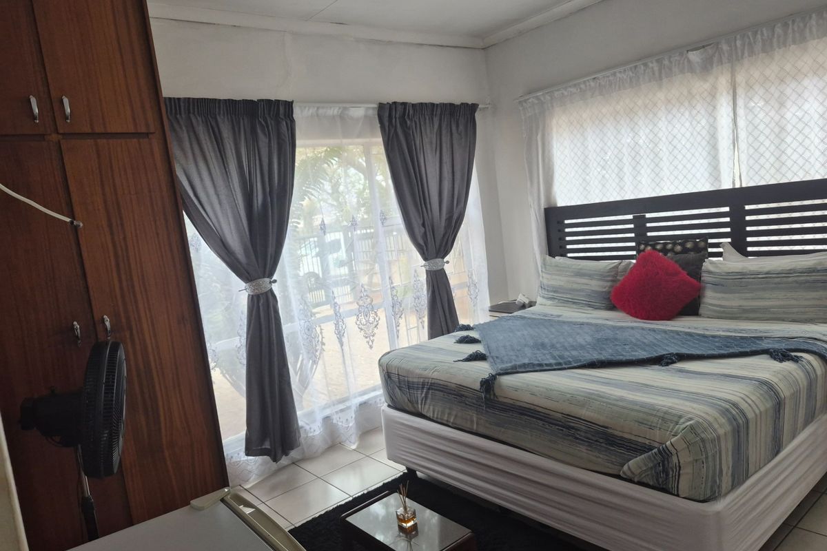 A bedroom featuring built-in wooden cupboards, tiled flooring, and large windows allowing natural light, with some visible wear on the ceiling finishes.