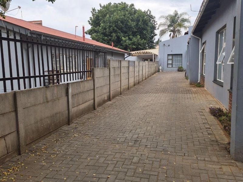 A paved side driveway or rear access path featuring a precast concrete boundary wall and metal security fencing alongside the house structure.
