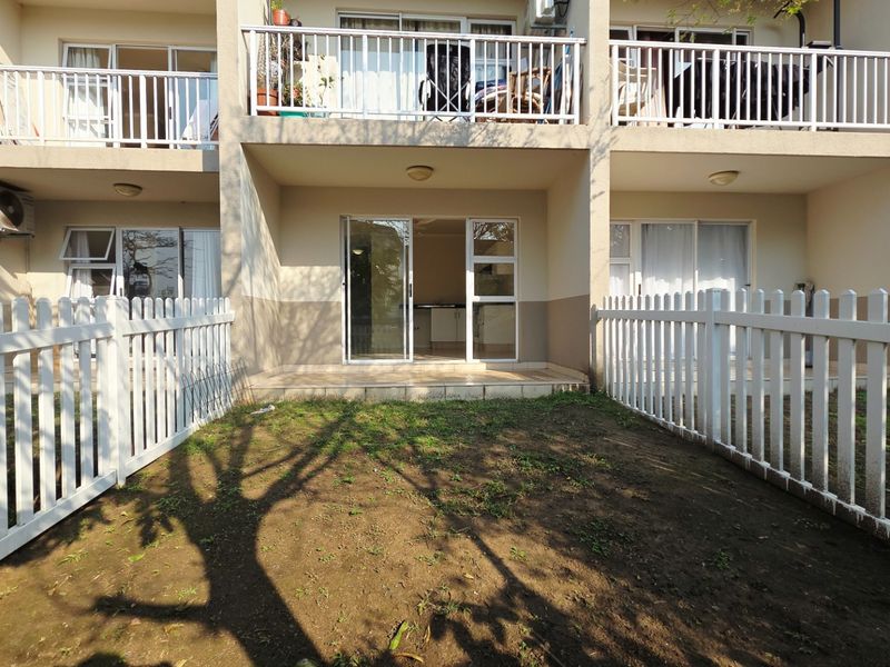 Rear view of an apartment unit featuring a private garden area with a white picket fence, covered patio, and sliding door access.