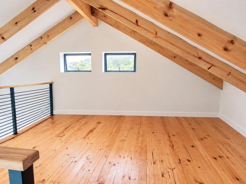 A bright, open-plan loft bedroom featuring exposed timber rafters, polished timber flooring, and a modern metal balustrade overlooking the lower level.