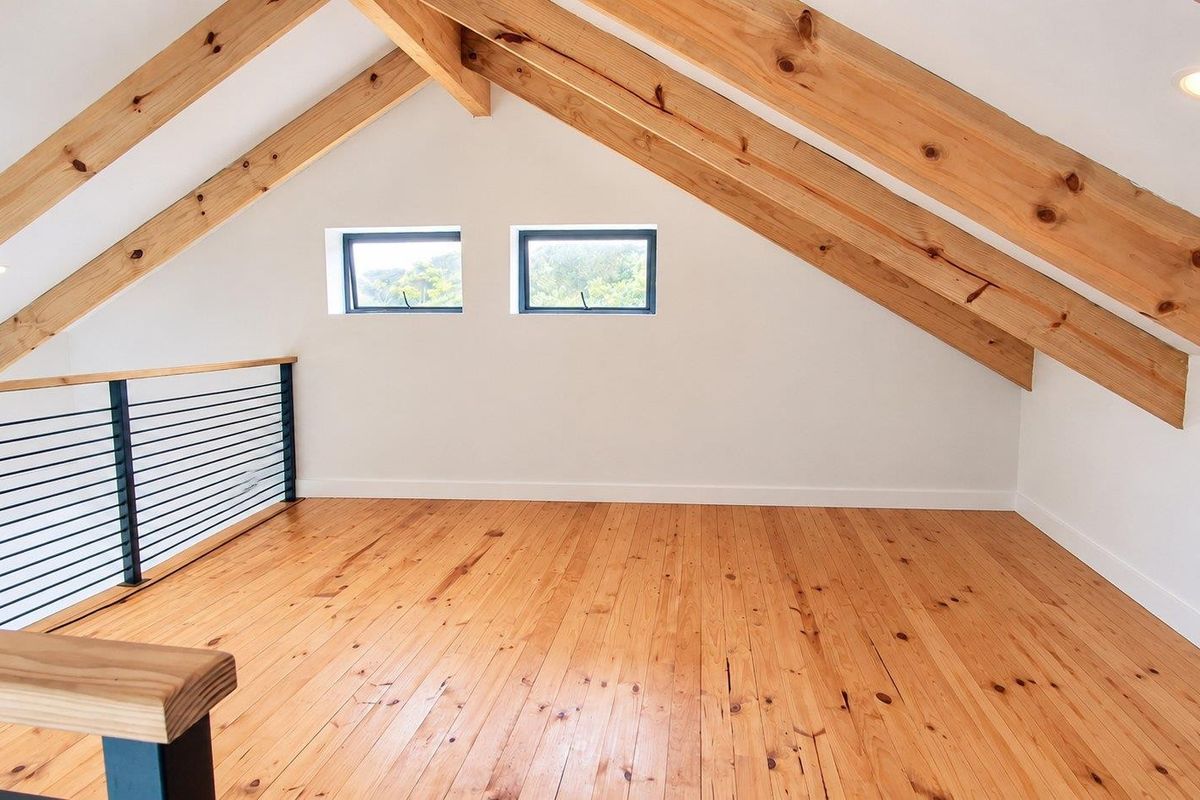A bright, open-plan loft bedroom featuring exposed timber rafters, polished timber flooring, and a modern metal balustrade overlooking the lower level.