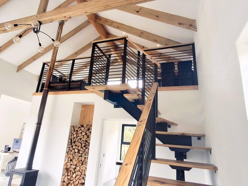 Spacious living area featuring high vaulted ceilings with exposed timber trusses, a modern mezzanine level, and a striking staircase with a built-in fireplace.