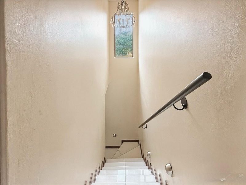 A bright, minimalist staircase featuring tiled steps, a sleek handrail, and a tall window that allows natural light to illuminate the hallway.