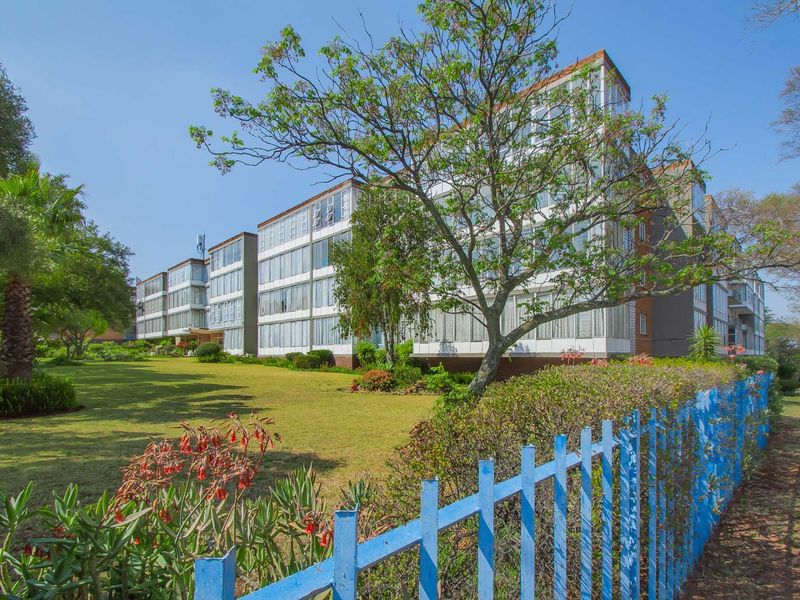 Exterior view of a multi-story apartment building featuring extensive glass facades, set within a well-maintained garden area with perimeter fencing.
