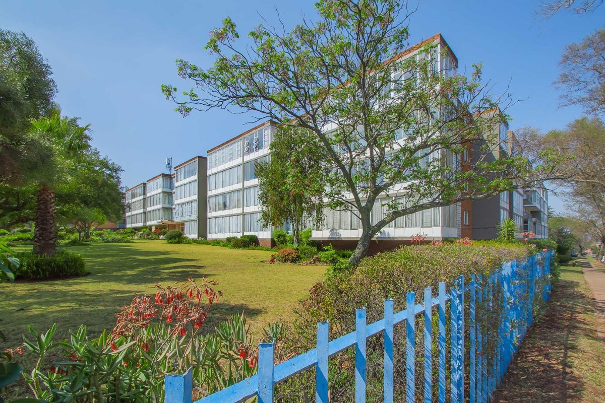Exterior view of a multi-story apartment building featuring extensive glass facades, set within a well-maintained garden area with perimeter fencing.