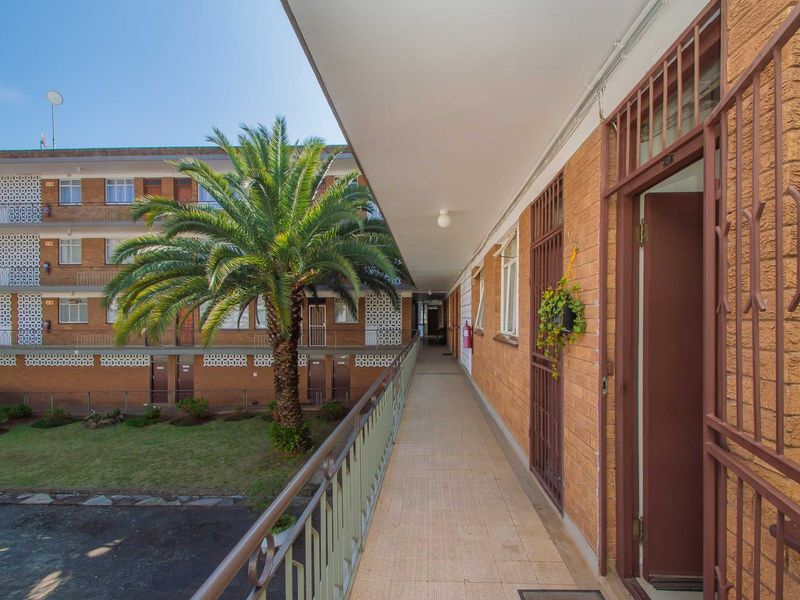 An exterior apartment walkway featuring tiled flooring, metal security gates, and a view overlooking the complex courtyard and garden area.